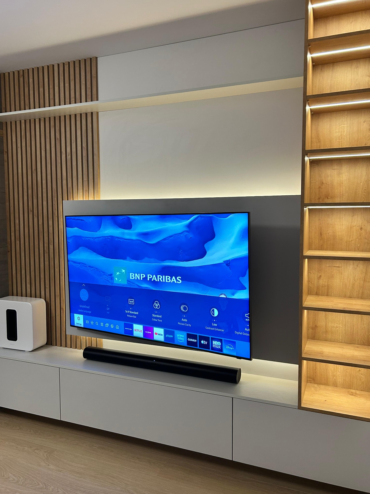 Nowoczesna zabudowa ściany z TV, z drewnianymi lamelami i podświetlanymi półkami. Minimalistyczny design, białe szafki dolne i szary panel za telewizorem.