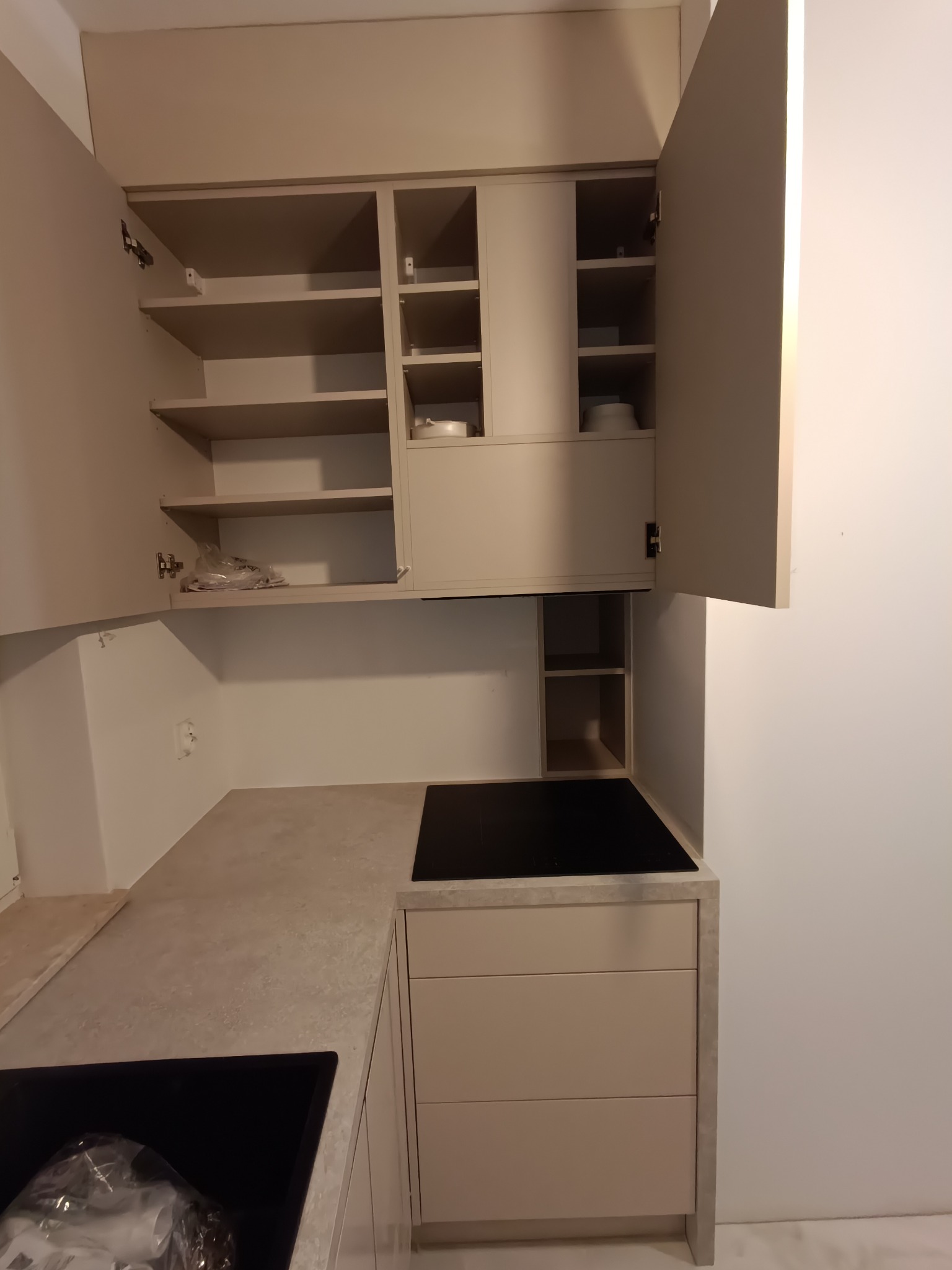 Jasna kuchnia z otwartymi szafkami, widoczna płyta indukcyjna i blat o teksturze betonu. Minimalistyczny design, ciepłe oświetlenie.