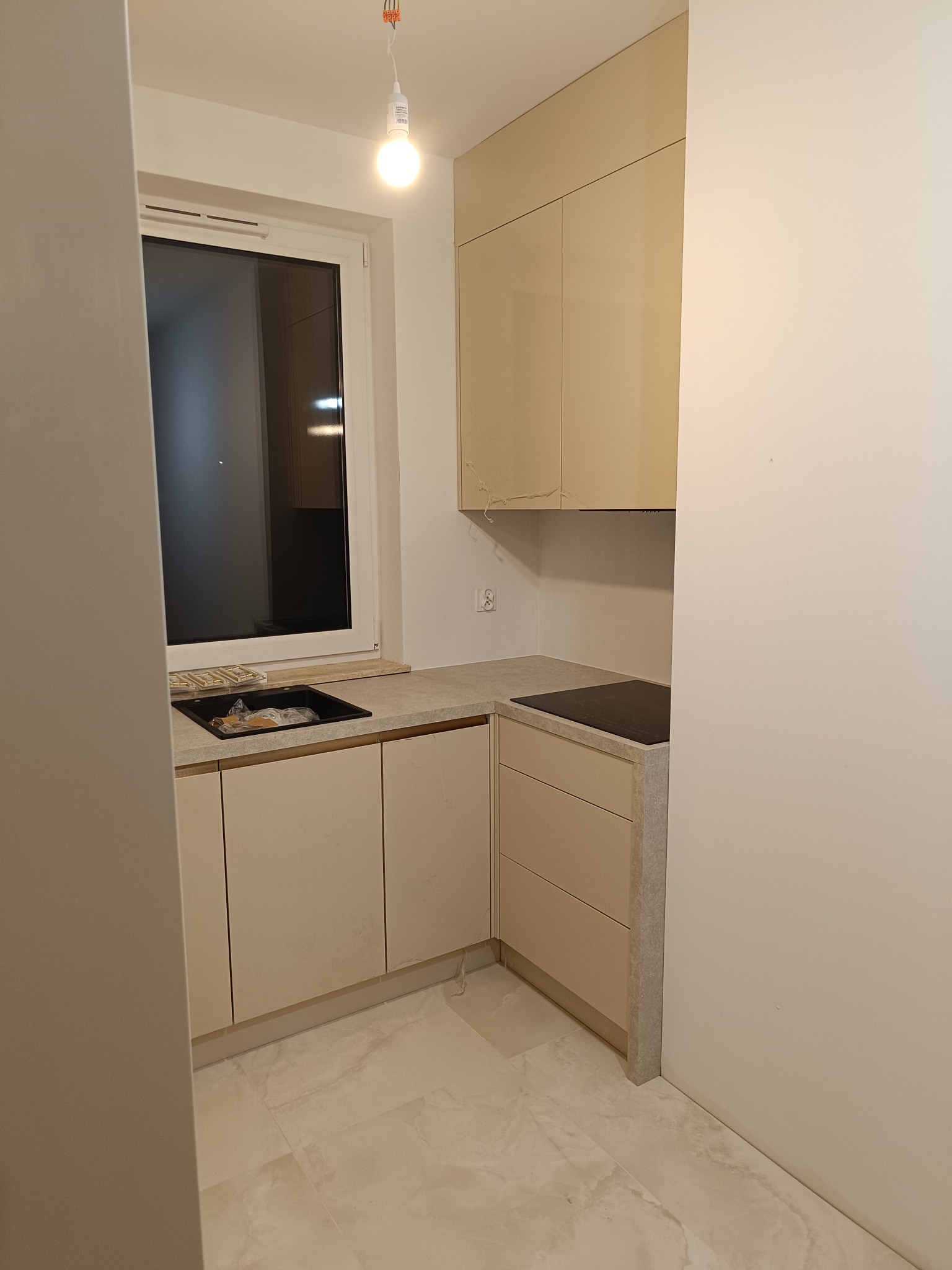 Jasna kuchnia na wymiar z beżowymi frontami i kamiennym blatem, widok na zlew i płytę indukcyjną. Minimalistyczny design, nowoczesne wykończenie.