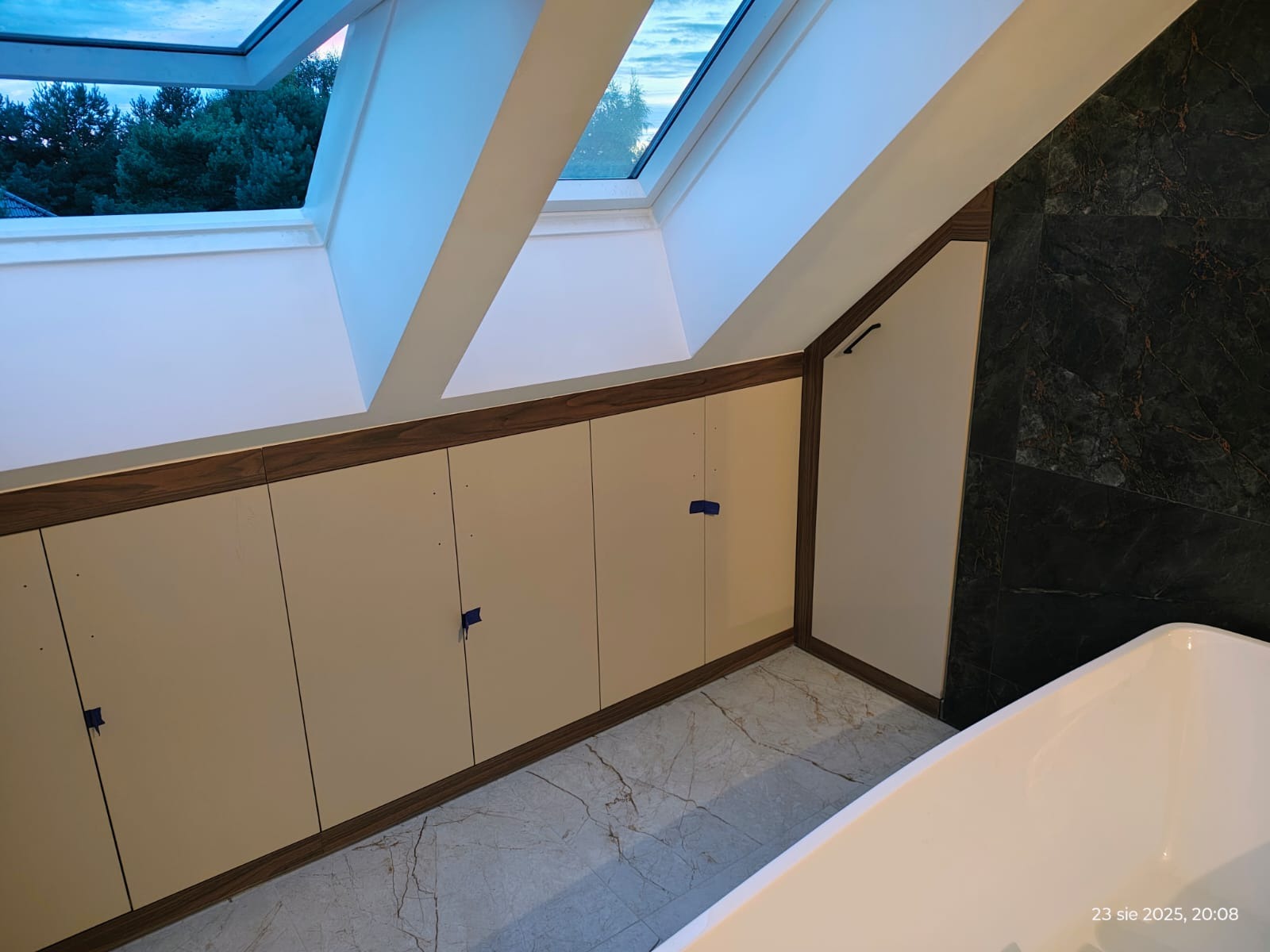 Szafki wnękowe z beżowymi frontami i ciemnobrązowym obramowaniem, zamontowane pod skosem dachu w łazience z marmurową podłogą i czarną ścianą.