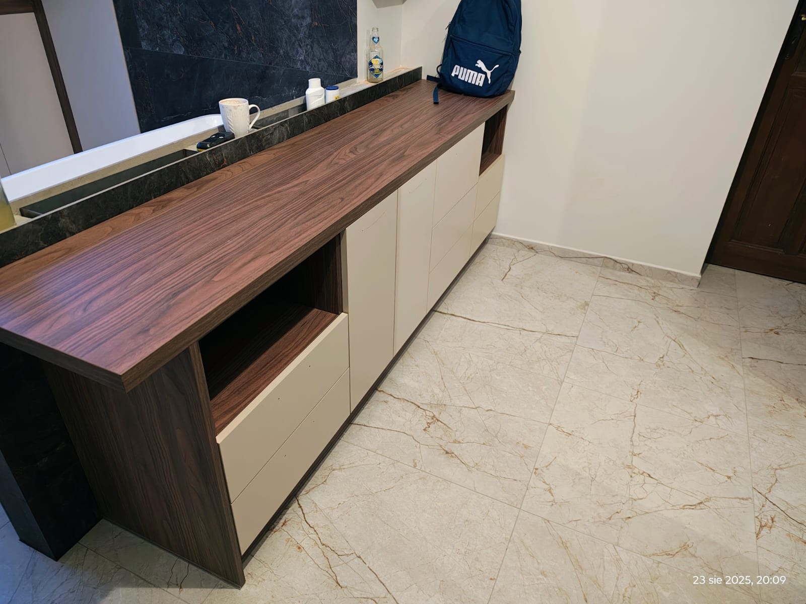 Nowoczesna szafka łazienkowa z blatem z drewna orzechowego i jasnymi frontami. Widoczna umywalka w tle i plecak Puma na blacie. Marmurowe płytki podłogowe.