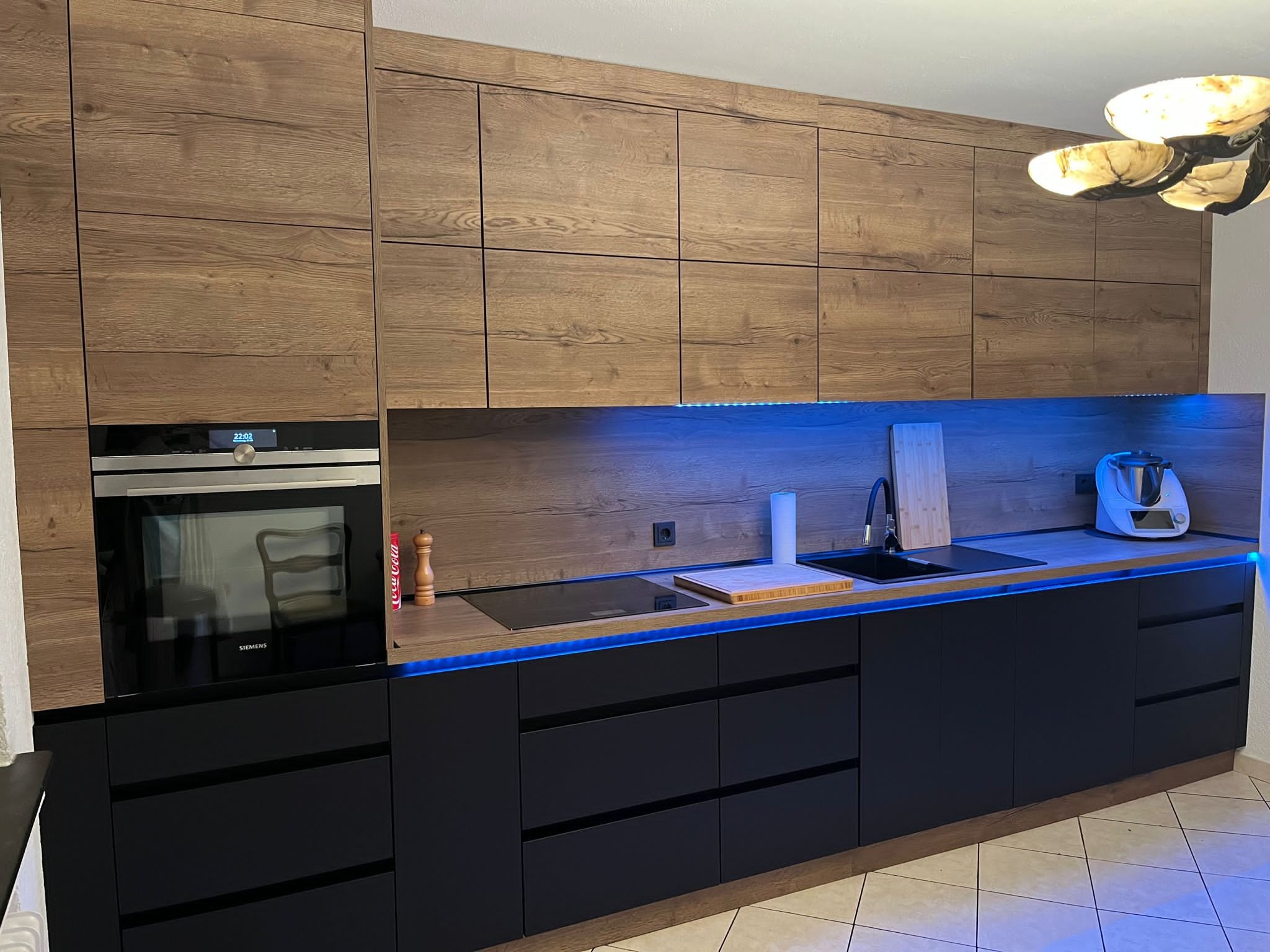 Nowoczesna kuchnia na wymiar: czarne szafki dolne, drewnopodobne górne, podświetlenie LED, piekarnik Siemens i sprzęt AGD na blacie. Stylowe oświetlenie sufitowe.
