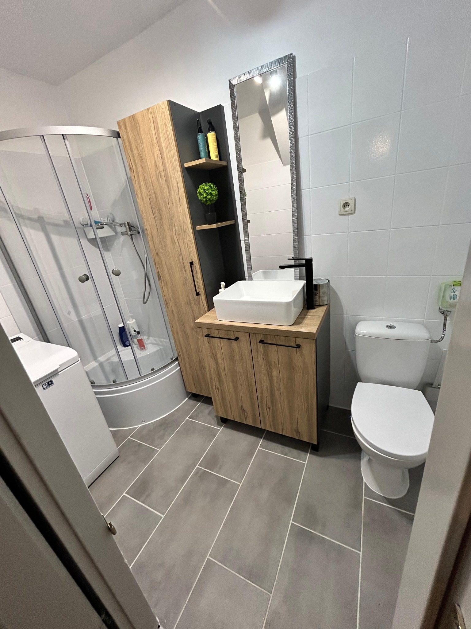 Nowoczesna łazienka z kabiną prysznicową, szafką z umywalką i wysoką szafką z półkami. Uwagę przykuwają szare płytki podłogowe i białe płytki ścienne.