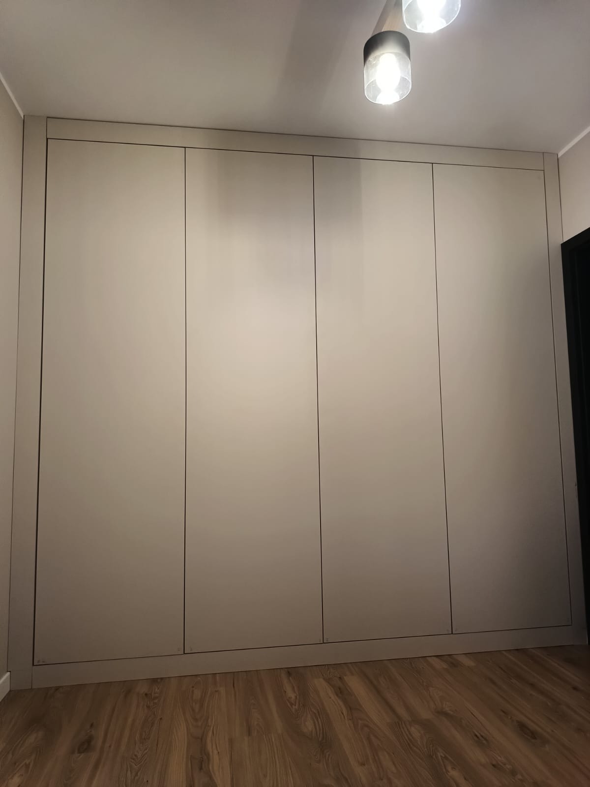 Czterodrzwiowa szafa wnękowa w odcieniach beżu, z minimalistycznymi uchwytami, dopasowana do wnętrza z drewnianą podłogą i nowoczesnym oświetleniem sufitowym.