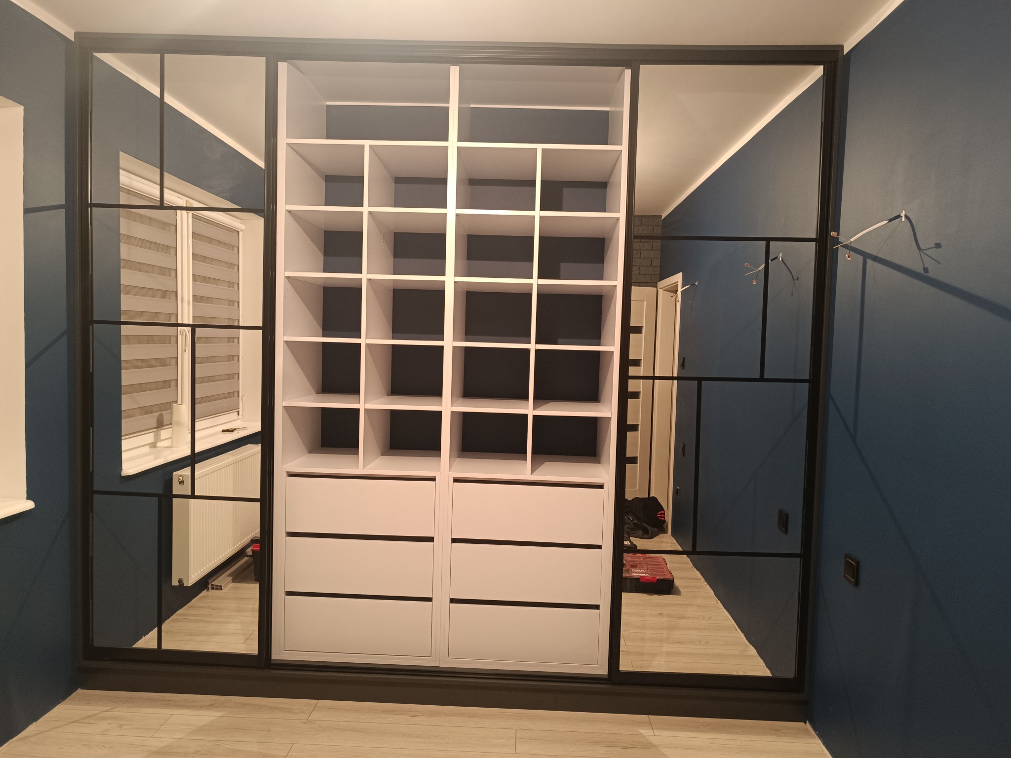 Biała szafa wnękowa z czarnymi ramami i lustrami w Barcinie. Widoczne półki i szuflady. Nowoczesny design, niebieska ściana w tle.