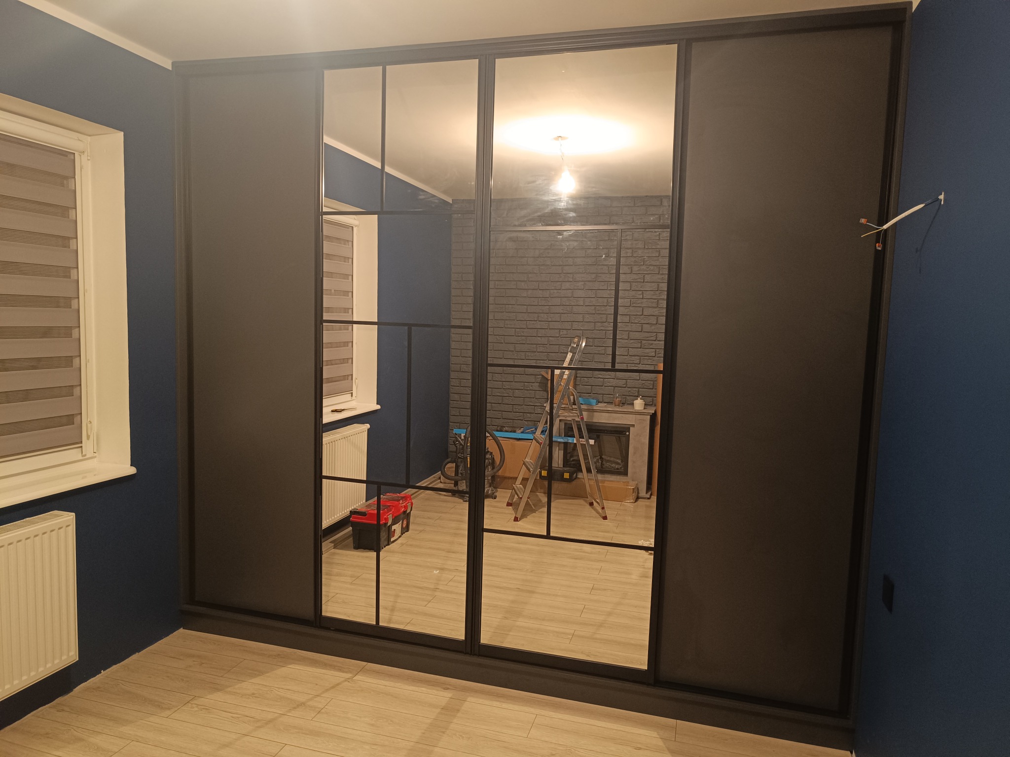 Nowoczesna szafa wnękowa z przesuwnymi drzwiami z czarnymi ramami i lustrami, odbijająca wnętrze pokoju z ceglaną ścianą i kominkiem.