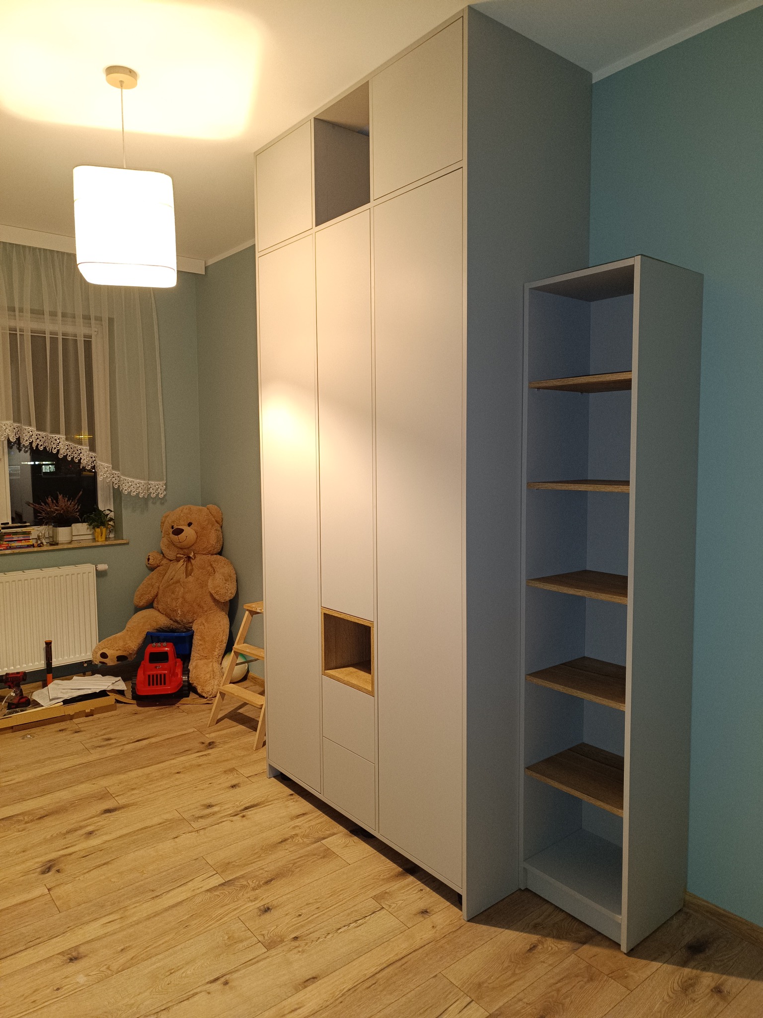 Jasnoszara szafa z półkami z drewna w pokoju dziecięcym, obok regał z półkami. Na podłodze zabawki, w tle okno z zasłonami i pluszowy miś.