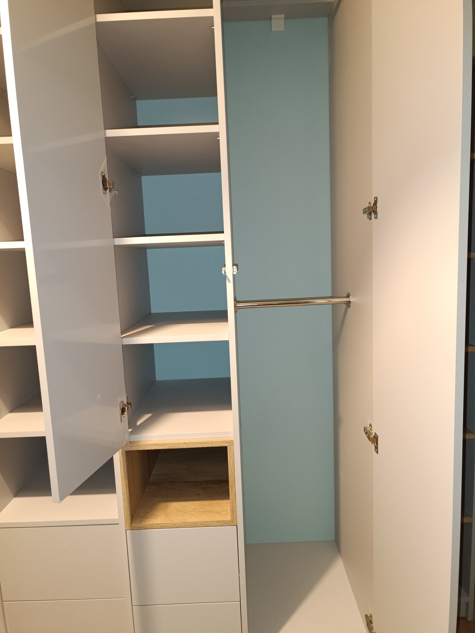 Biała szafa wnękowa z otwartymi drzwiami, widoczne półki i drążek na ubrania. Jedna z półek w kolorze drewna. Minimalistyczny design, jasne wnętrze.