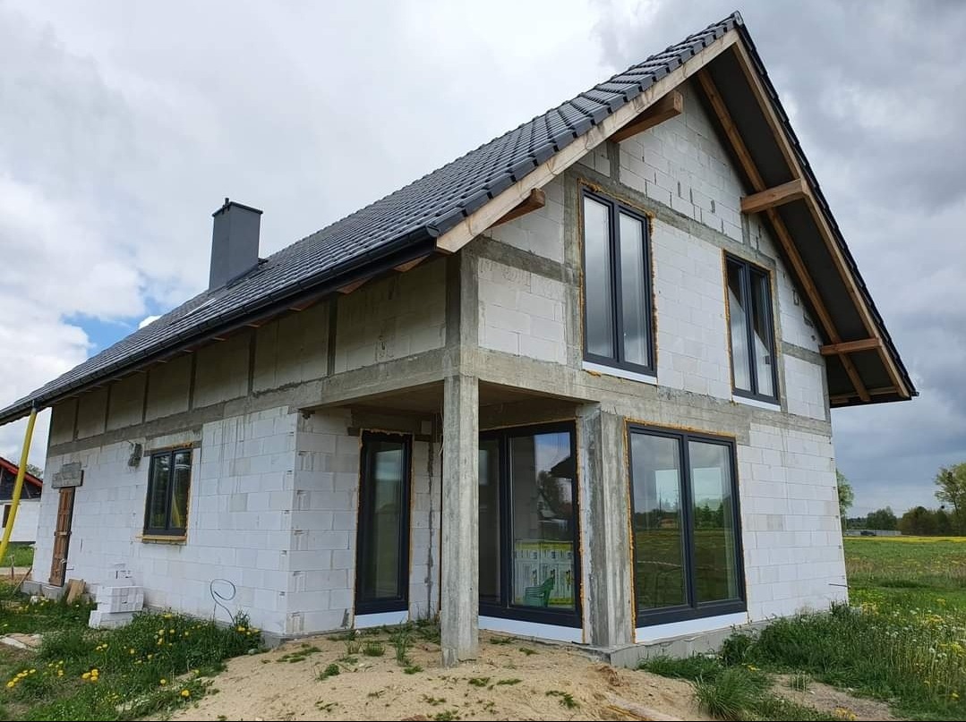 Dom w trakcie budowy z zamontowanymi oknami w grafitowych ramach. Widoczne bloczki betonowe i elementy konstrukcji dachu. Tło stanowi łąka i pochmurne niebo.