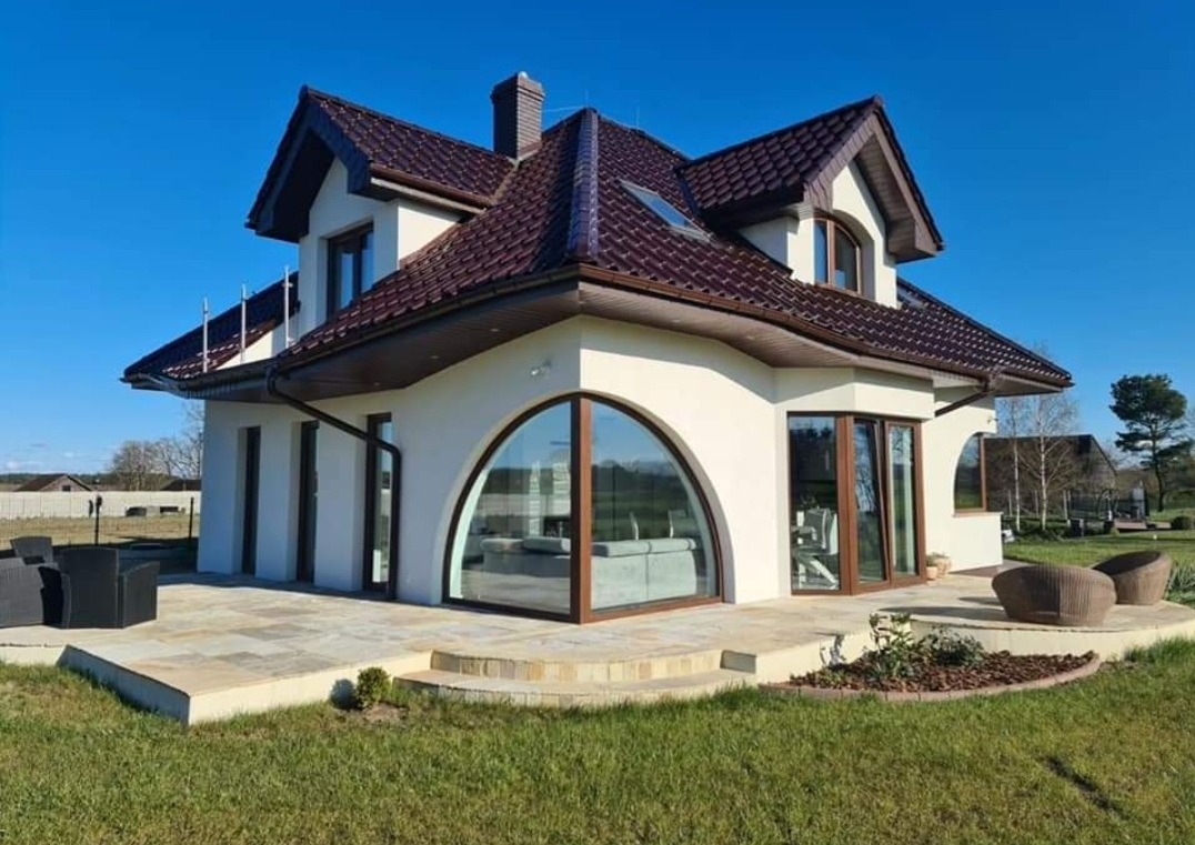 Elegancki dom z kremową elewacją, brązowym dachem i dużym, łukowym oknem. Widoczny taras z meblami ogrodowymi i zadbany trawnik. Błękitne niebo w tle.