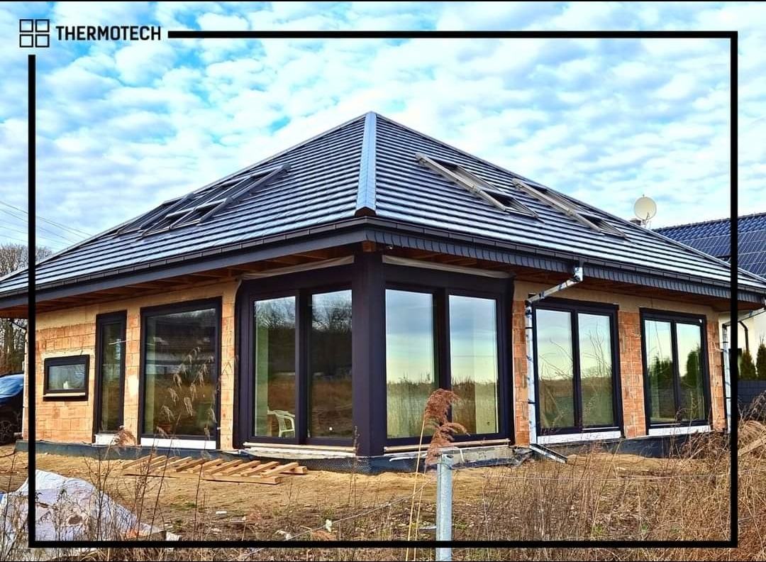 Dom w trakcie budowy z zamontowanymi oknami i drzwiami przesuwnymi w ciemnych ramach. Widoczne surowe ściany z cegły i dach z oknami dachowymi.