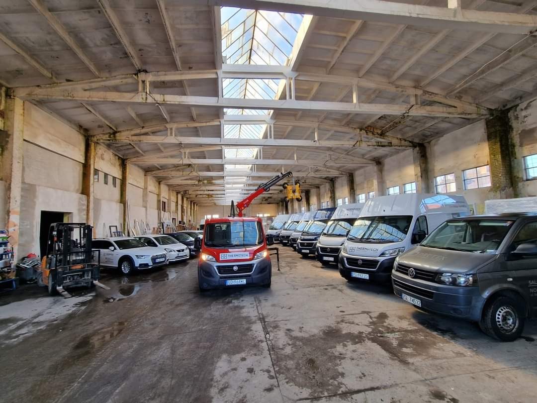 Flota pojazdów firmy z podnośnikiem w hali magazynowej z oknami dachowymi. Widoczny wózek widłowy i samochody osobowe. Logo firmy na pojazdach.