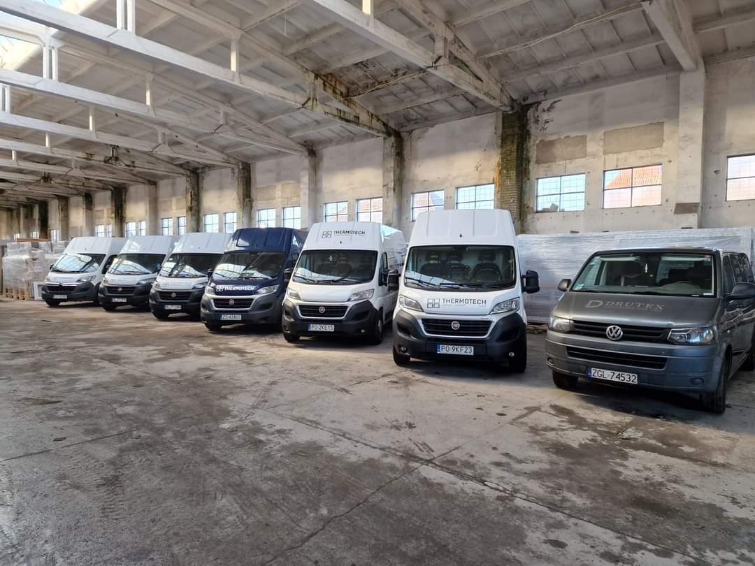Flota firmowa: równe rzędy busów Thermotech i Drutex w hali magazynowej, gotowe do transportu okien i drzwi. Jasne światło podkreśla czystość i profesjonalizm.