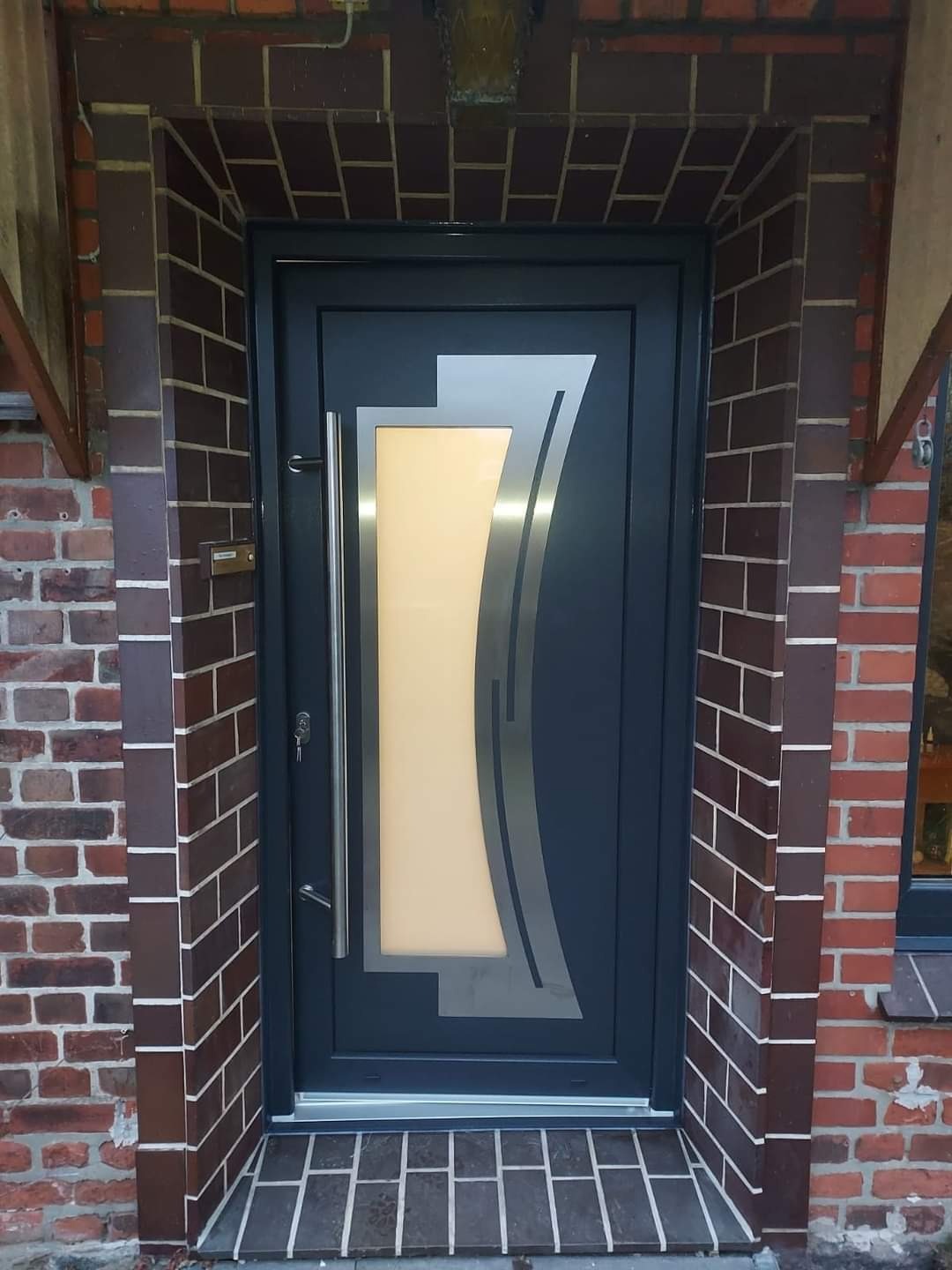 Grafitowe drzwi wejściowe z szybą i srebrnymi zdobieniami, zamontowane w murze z cegły. Długi, pionowy uchwyt ze stali nierdzewnej. Widoczny zamek i skrzynka na listy.