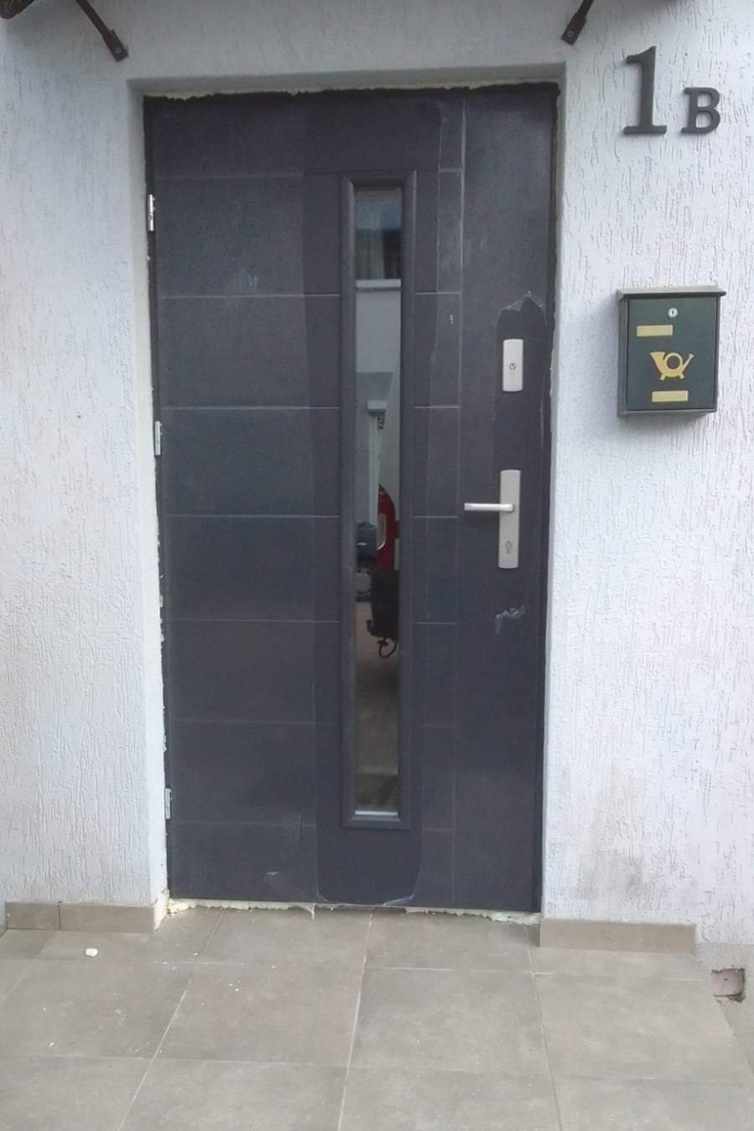 Grafitowe drzwi wejściowe z szybą, zamontowane w jasnym budynku z numerem 1B i skrzynką pocztową. Widoczne ślady pianki montażowej.