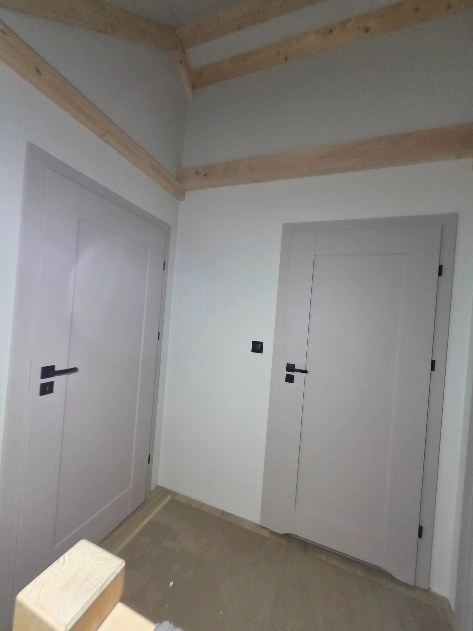 Dwa białe, minimalistyczne drzwi z czarnymi klamkami w jasnym wnętrzu z drewnianymi belkami stropowymi. Ujęcie z niskiej perspektywy.