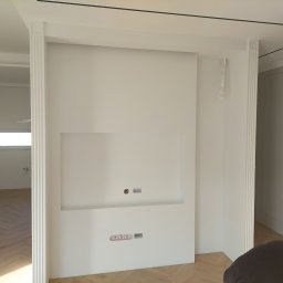 REMAR MARIUSZ GODLEWSKI - Biała ściana z wnęką na TV i gniazdkami, wykończona sztukaterią. Widoczny fragment podłogi z parkietem w jodełkę i fragment osoby w prawym dolnym rogu. Minimalistyczny design.