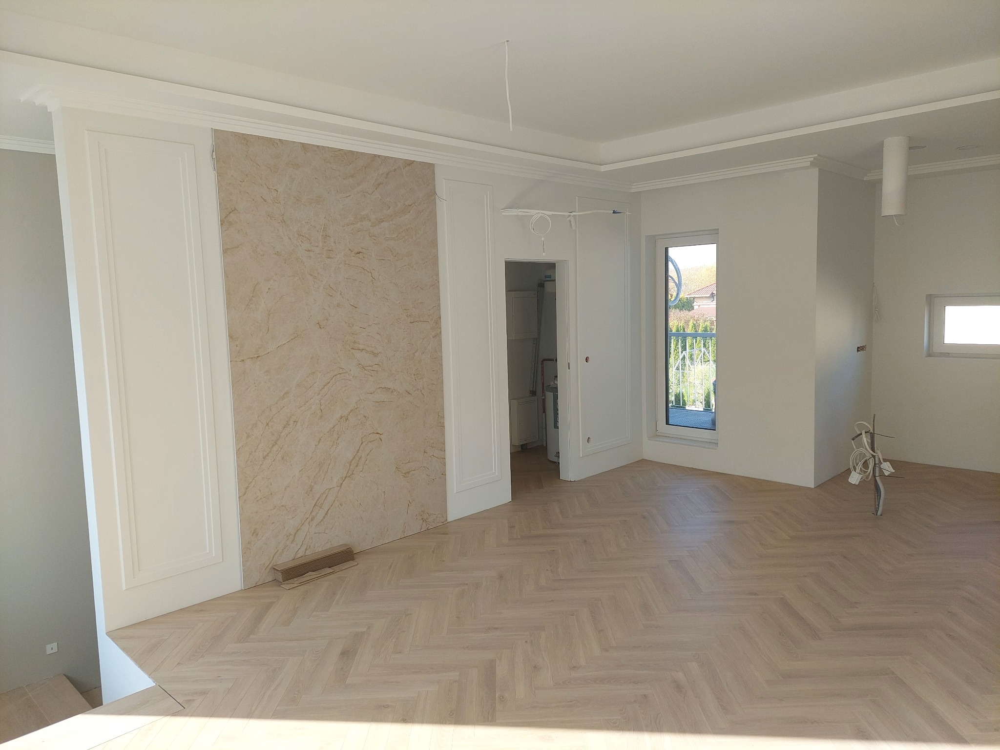 Jasne, przestronne wnętrze po remoncie z parkietem w jodełkę, dekoracyjnymi panelami ściennymi i sztukaterią sufitową. Widoczne okno balkonowe i częściowo wyposażone pomieszczenie.