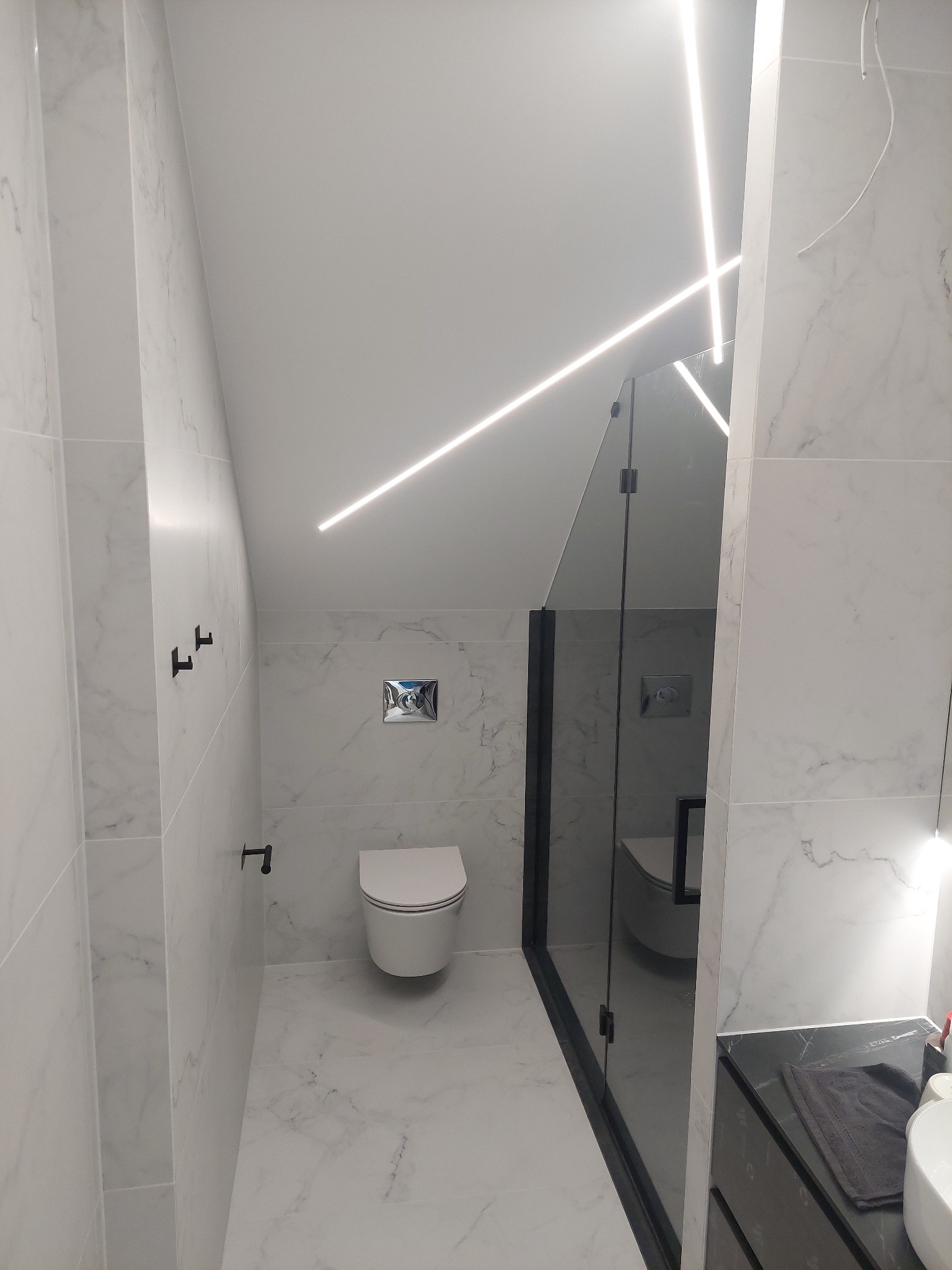 Nowoczesna łazienka na poddaszu z białymi marmurowymi płytkami, kabiną prysznicową typu walk-in z czarnymi ramami i liniowym oświetleniem LED na suficie.