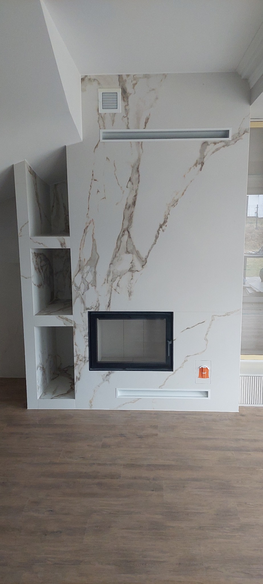 Wykończona ściana kominkowa z imitacją marmuru, wnękami i kratkami wentylacyjnymi. Nowoczesny design z drewnianą podłogą.