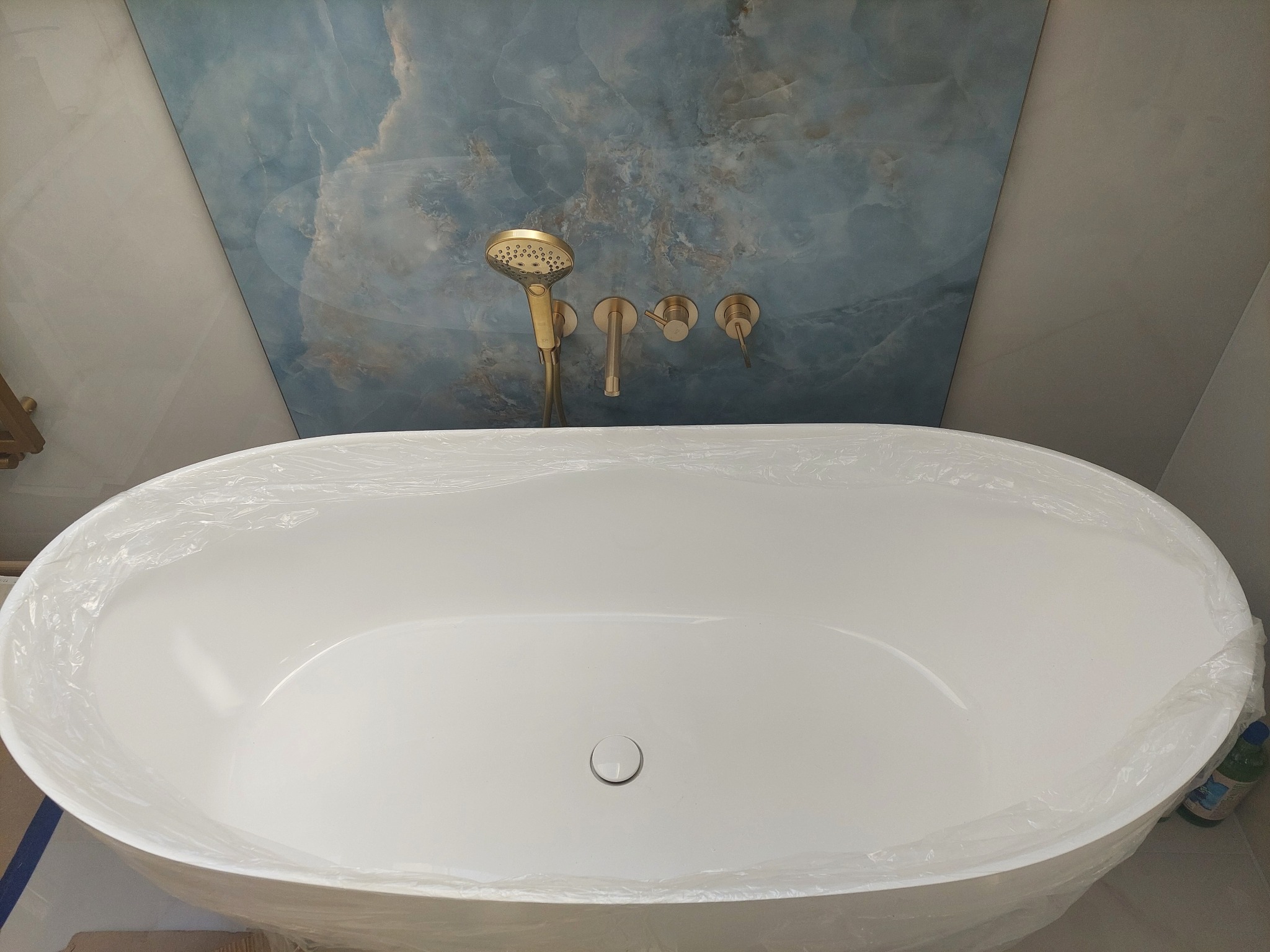 Biała wanna wolnostojąca z złotą armaturą na tle ściany z niebieskiego marmuru. Wanna zabezpieczona folią ochronną. Widok z góry.