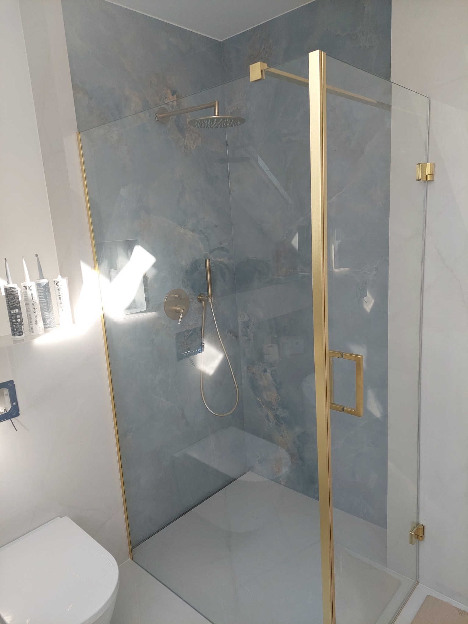 Nowoczesna kabina prysznicowa z złotymi profilami i deszczownicą na tle ściany z efektem marmuru. Widoczne silikony i toaleta. Wykończenie łazienki w detalu.