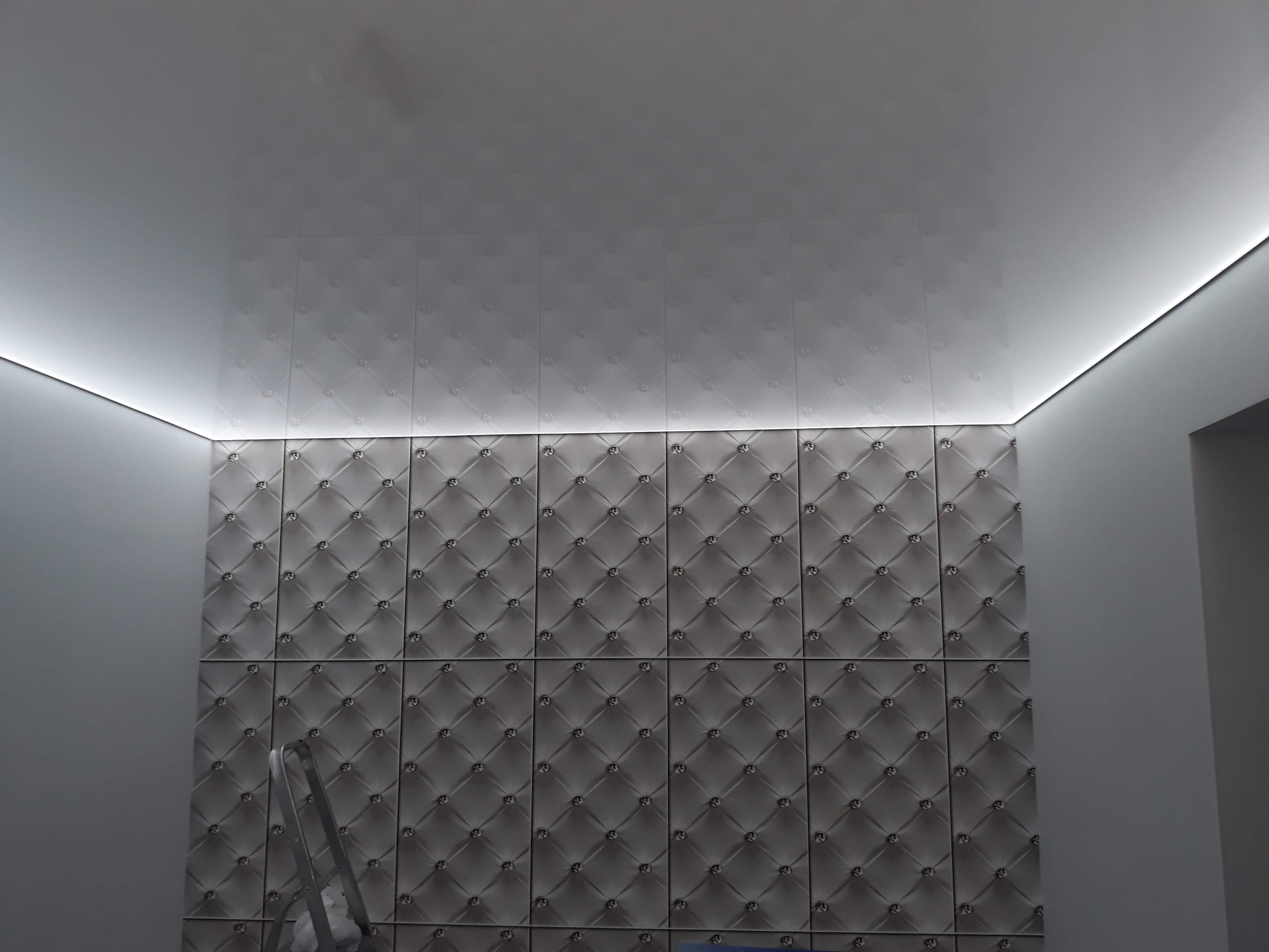 Panel dekoracyjny 3D na ścianie z oświetleniem LED podsufitowym. Widoczny fragment drabiny malarskiej. Eleganckie wykończenie wnętrza w odcieniach szarości.