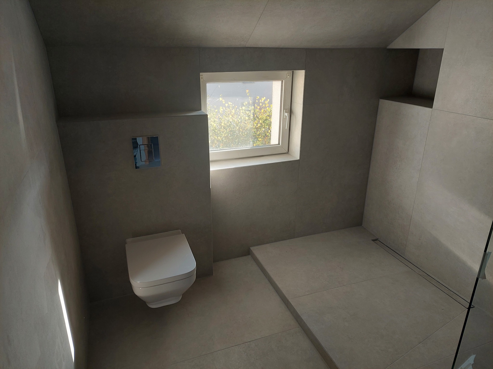 Minimalistyczna łazienka w odcieniach szarości z podwieszaną toaletą, oknem i odpływem liniowym w strefie prysznicowej. Nowoczesne wykończenie z płytek wielkoformatowych.