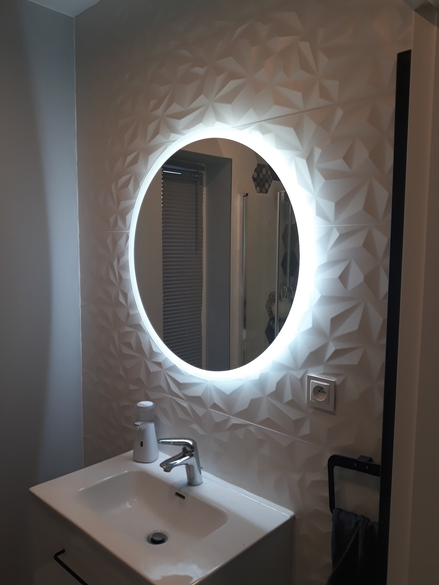 Nowoczesna łazienka z umywalką, baterią i okrągłym lustrem z podświetleniem LED na ścianie z trójwymiarowymi płytkami. Widoczny dozownik mydła i wieszak na ręcznik.