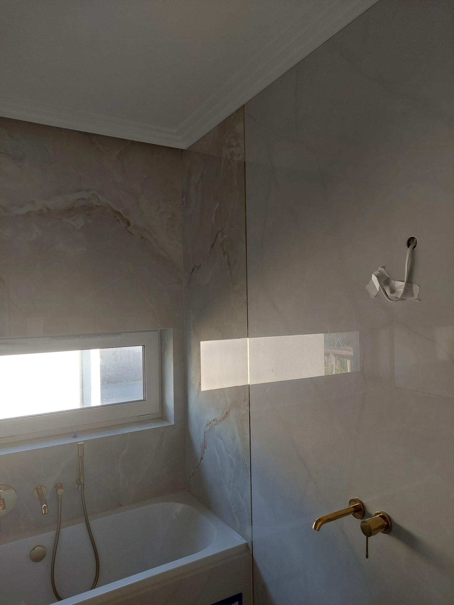 Wykończona łazienka z marmurowymi płytkami, złotą armaturą i oknem. Widoczne detale wykończeniowe i estetyczne połączenie materiałów.