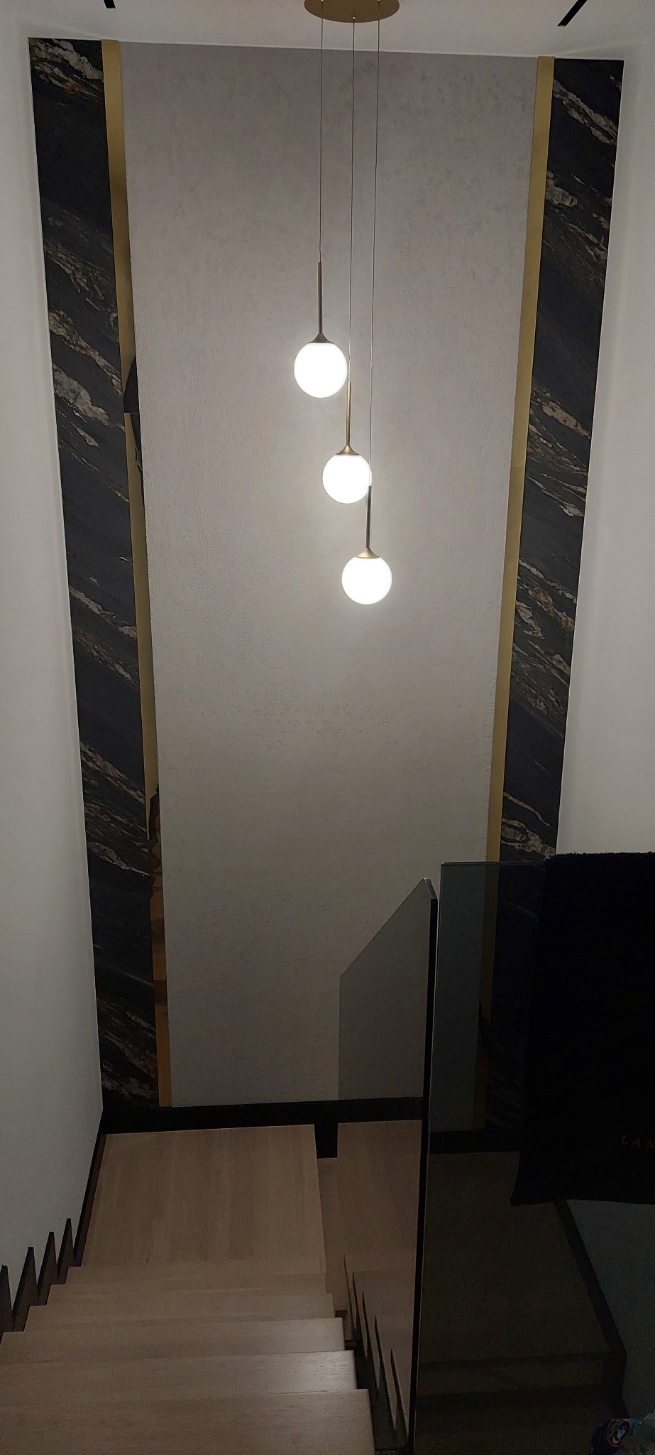 Nowoczesne wykończenie klatki schodowej z marmurowymi panelami, złotymi akcentami i designerską lampą wiszącą z trzema kulami. Widok z góry na drewniane schody.