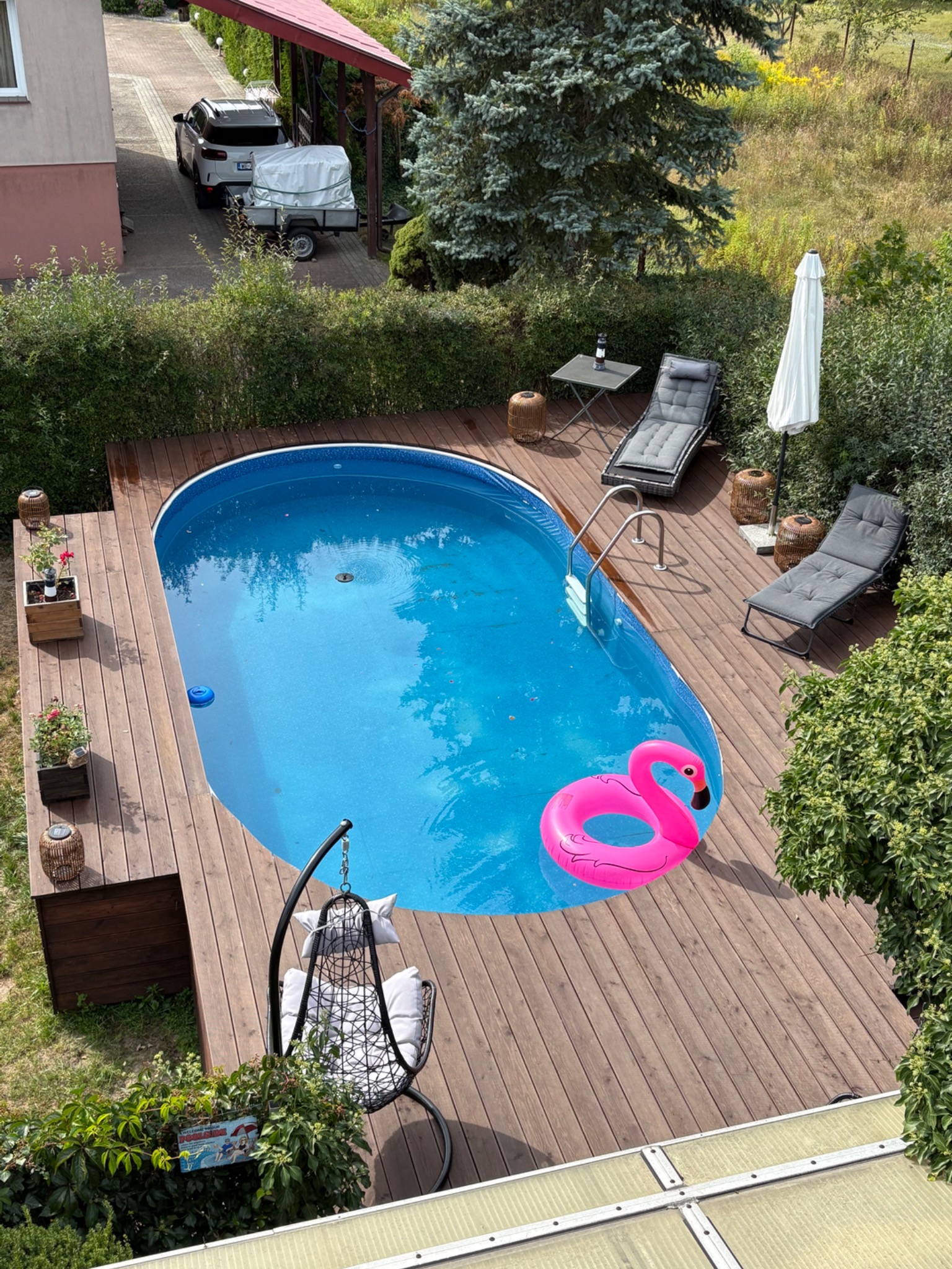 Owalny basen z drewnianym tarasem, leżakami i dmuchanym flamingiem. Widok z góry na relaksujący ogród z elementami dekoracyjnymi i zielenią.