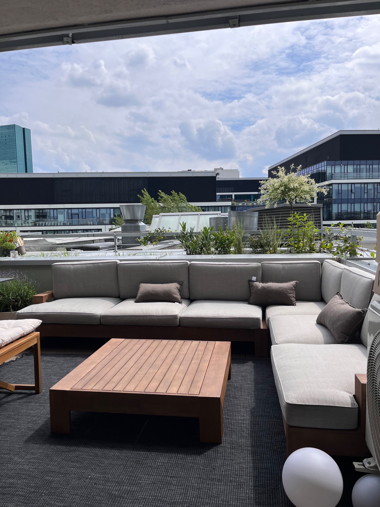 Nowoczesny taras na dachu z szarą sofą narożną, drewnianym stolikiem kawowym i zielenią w donicach. W tle budynki biurowe i błękitne niebo z chmurami.