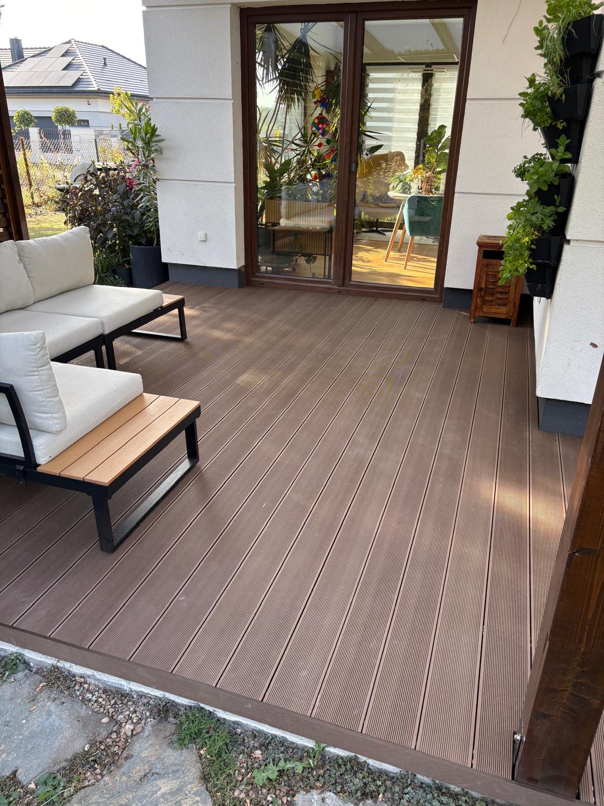 Nowoczesny taras z deski kompozytowej w odcieniach brązu, z jasnymi meblami ogrodowymi i zielonymi roślinami wertykalnymi. Widoczne przesuwne drzwi tarasowe.