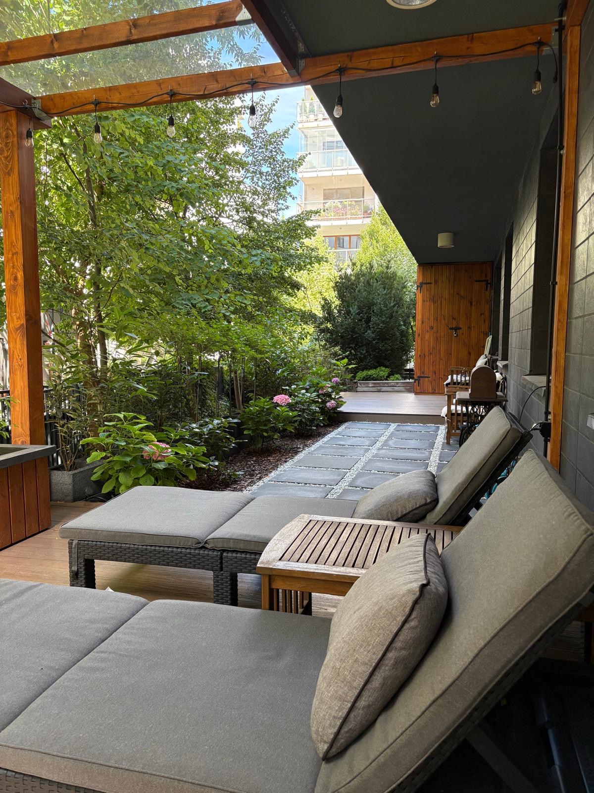 Zacieniony taras z leżakami i widokiem na bujny ogród z kwitnącymi hortensjami i ścieżką z kamiennych płyt. Oświetlenie girlandami żarówek.