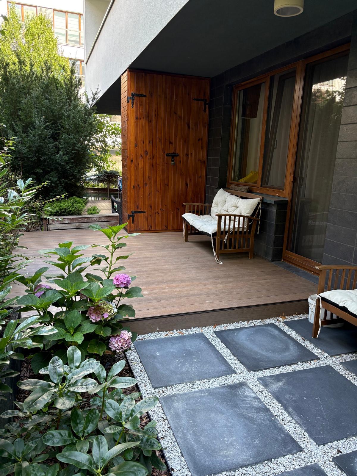 Nowoczesny taras z drewnianą podłogą, meblami ogrodowymi i kwitnącymi hortensjami. Kamienne płyty oddzielone białym żwirem tworzą ścieżkę.