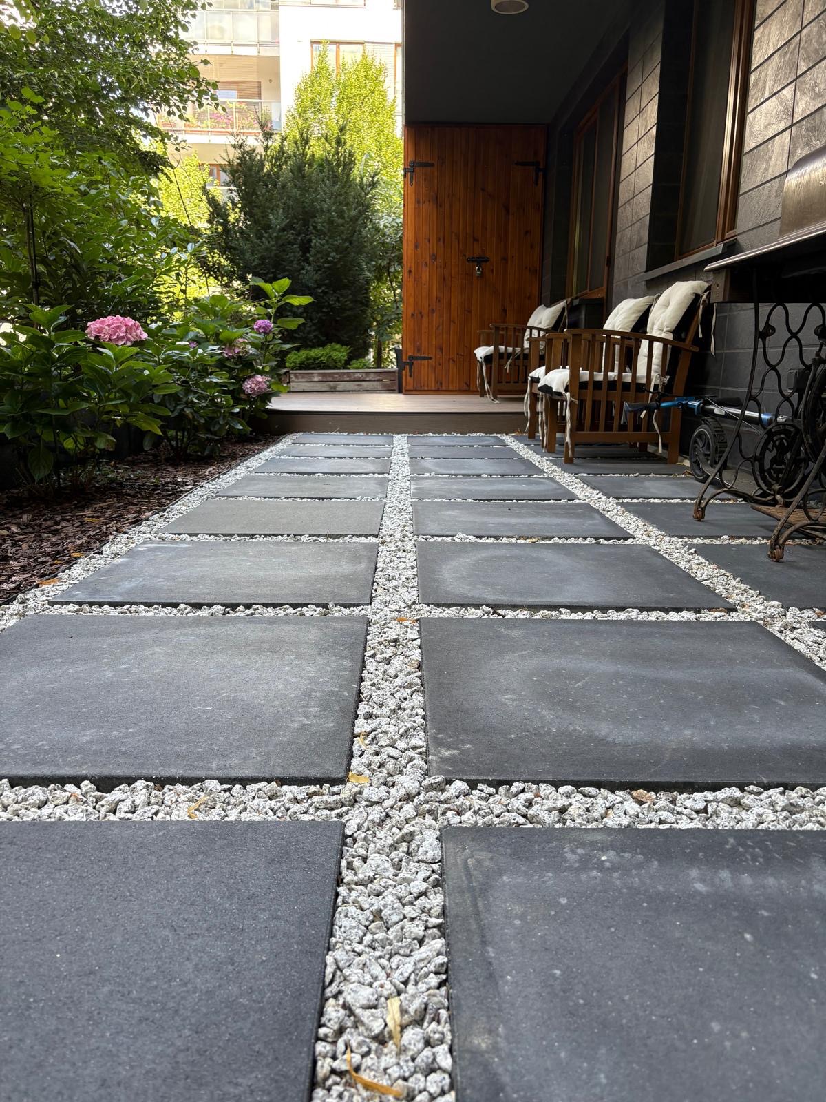 Ścieżka z szarych płyt chodnikowych oddzielonych białym żwirem, prowadząca do drewnianego ganku z krzesłami i zielenią w tle. Ujęcie z niskiej perspektywy.