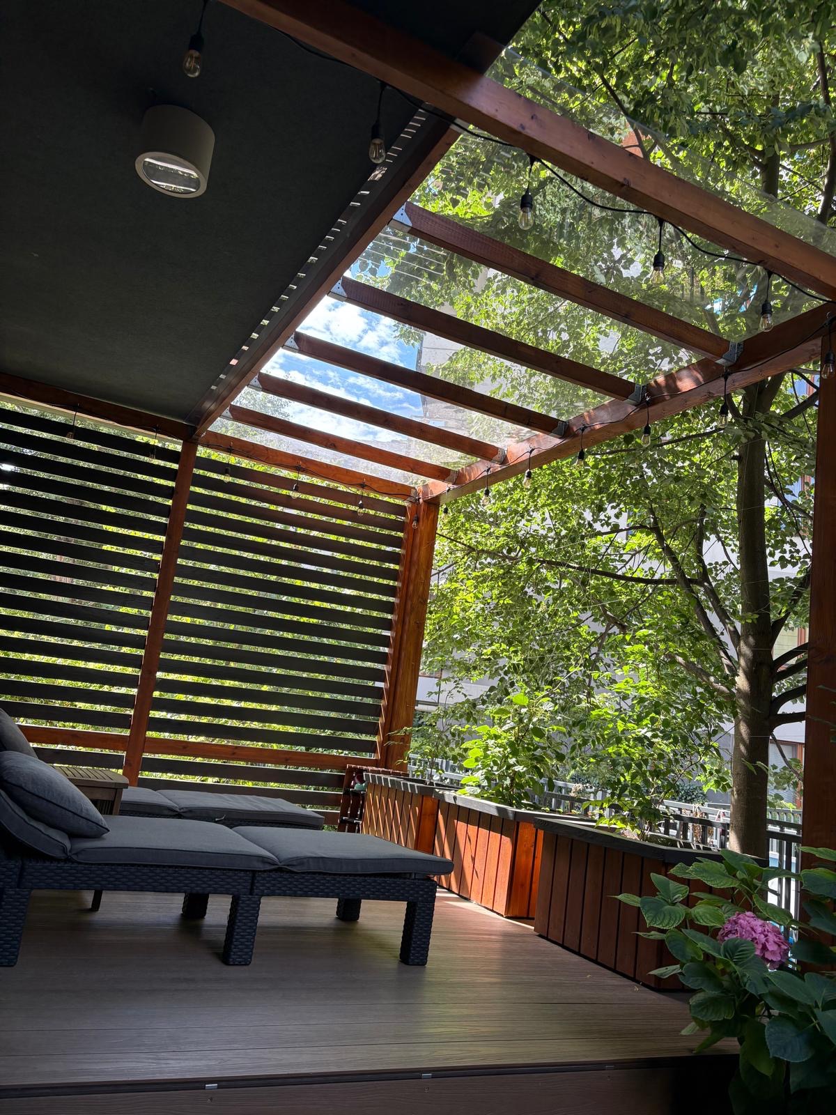 Zadaszony taras z drewnianą pergolą, szklanym dachem i lamelami, oświetlony girlandą żarówek. W tle zieleń drzew i błękit nieba. Wygodne leżaki z poduszkami.