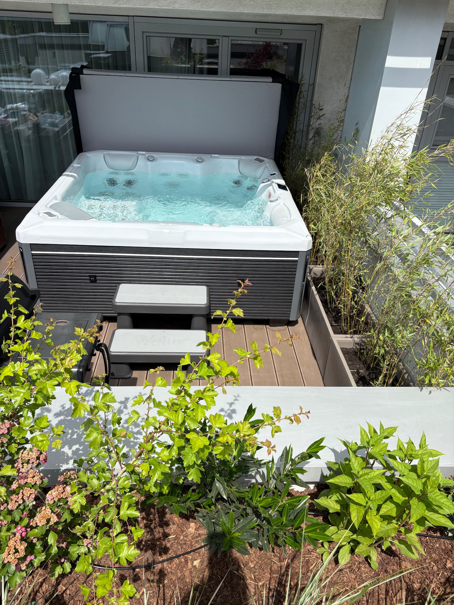 Nowoczesne jacuzzi na tarasie z drewnianą podłogą, otoczone zielenią w donicach. Widoczne schodki wejściowe i częściowo odsłonięta pokrywa. Jasne, naturalne światło.