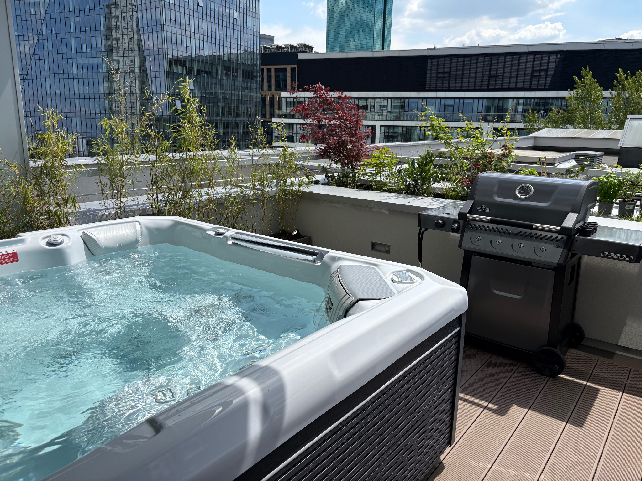 Nowoczesny taras na dachu z jacuzzi wypełnionym wodą i grillem gazowym, otoczony zielenią i wysokimi budynkami w tle. Słoneczny dzień.