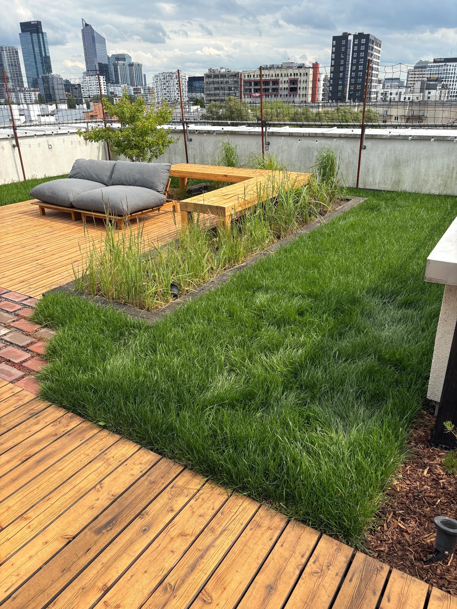 Nowoczesny ogród na dachu z drewnianym tarasem, sofą i zielonym trawnikiem na tle panoramy miasta. Unikalny układ roślin i oświetlenie dodają uroku.