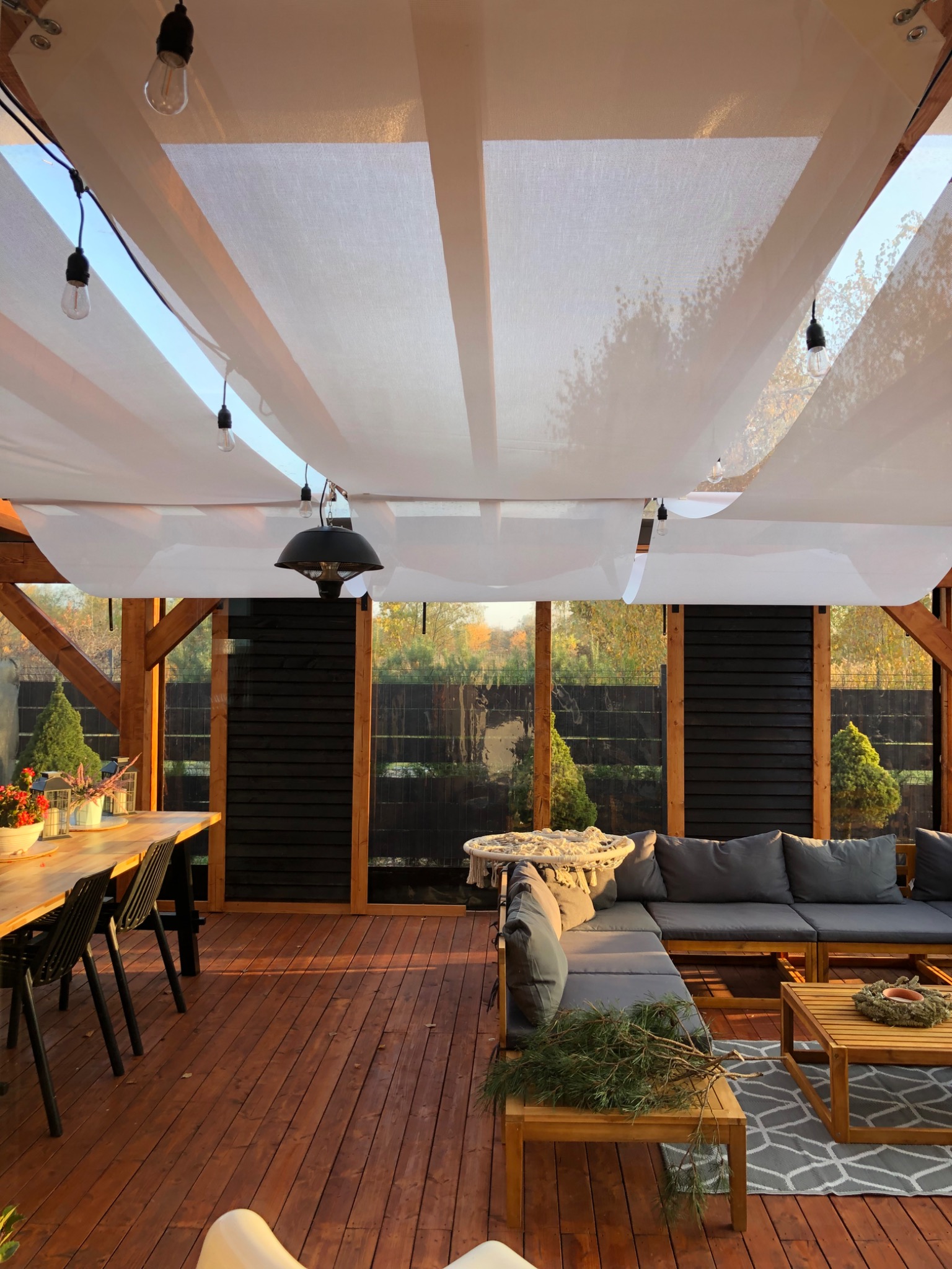 Zadaszony taras z drewnianą podłogą i meblami ogrodowymi, oświetlony wiszącymi żarówkami. Widoczne rośliny w tle i dekoracje na stole. Ciepłe światło zachodzącego słońca.