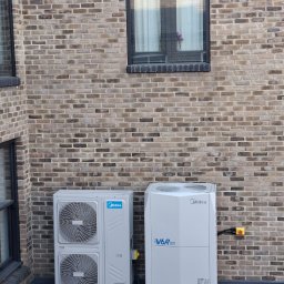 Mateusz Struzik MSAIR - Dwie jednostki klimatyzacji Midea V6R HEAT na tle ceglanej ściany budynku z oknami. Widoczne przewody i elementy montażowe. Ujęcie z dołu.