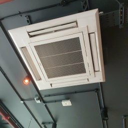 Mateusz Struzik MSAIR - Kasetonowa klimatyzacja sufitowa w szarej przestrzeni biurowej, widok z dołu. Widoczne czarne rury instalacyjne i oświetlenie punktowe.