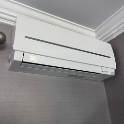 Mateusz Struzik MSAIR - Biały klimatyzator Mitsubishi Electric zamontowany na szarej ścianie w pomieszczeniu z dekoracyjną listwą sufitową. Widok z dołu, lekki kąt.