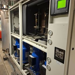 Mateusz Struzik MSAIR - Agregat chłodniczy Trane z zielonym wyświetlaczem, manometrami i niebieskimi sprężarkami. Widoczne rury i elementy instalacji. Całość w metalowej obudowie.