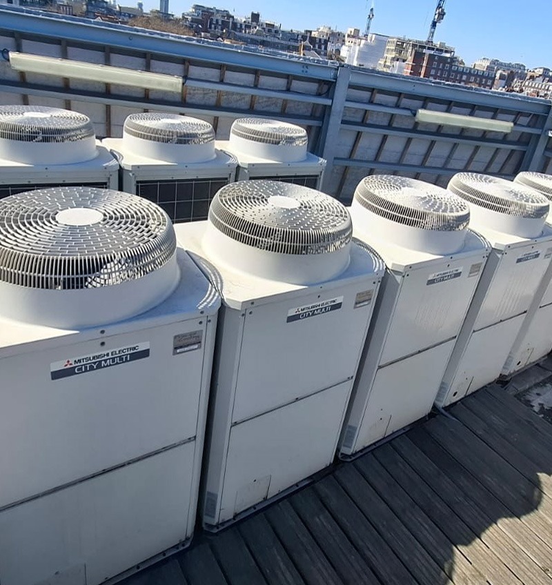 Rząd białych jednostek zewnętrznych klimatyzacji Mitsubishi Electric City Multi na dachu budynku, widoczne wentylatory i etykiety. W tle zabudowa miejska i błękitne niebo.