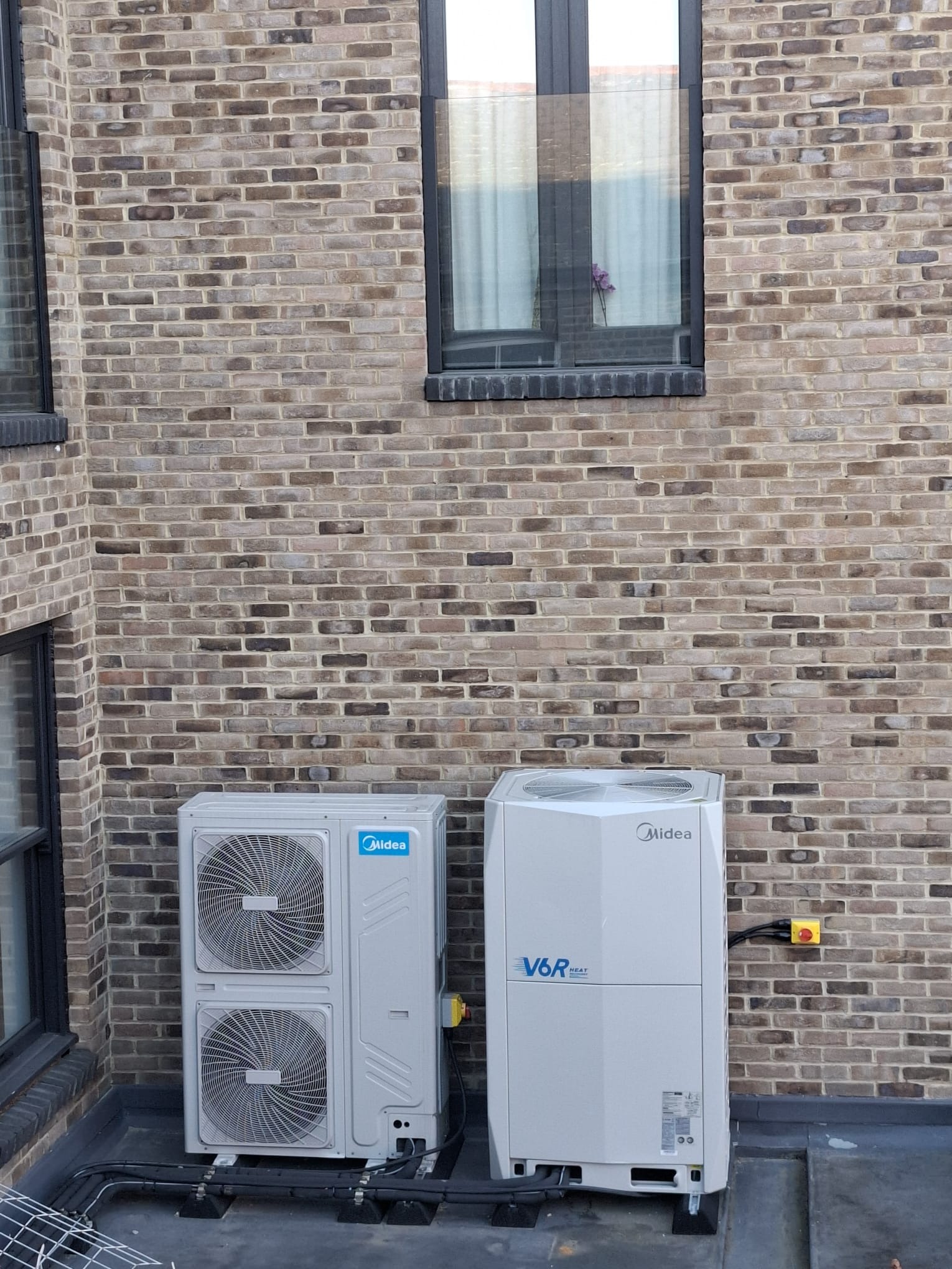 Dwie jednostki klimatyzacji Midea V6R HEAT na tle ceglanej ściany budynku z oknami. Widoczne przewody i elementy montażowe. Ujęcie z dołu.