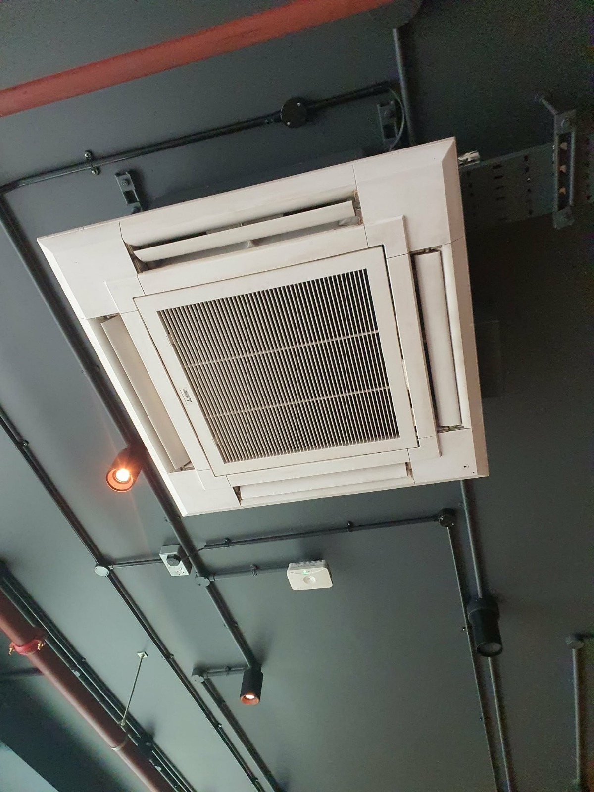 Kasetonowa klimatyzacja sufitowa w szarej przestrzeni biurowej, widok z dołu. Widoczne czarne rury instalacyjne i oświetlenie punktowe.