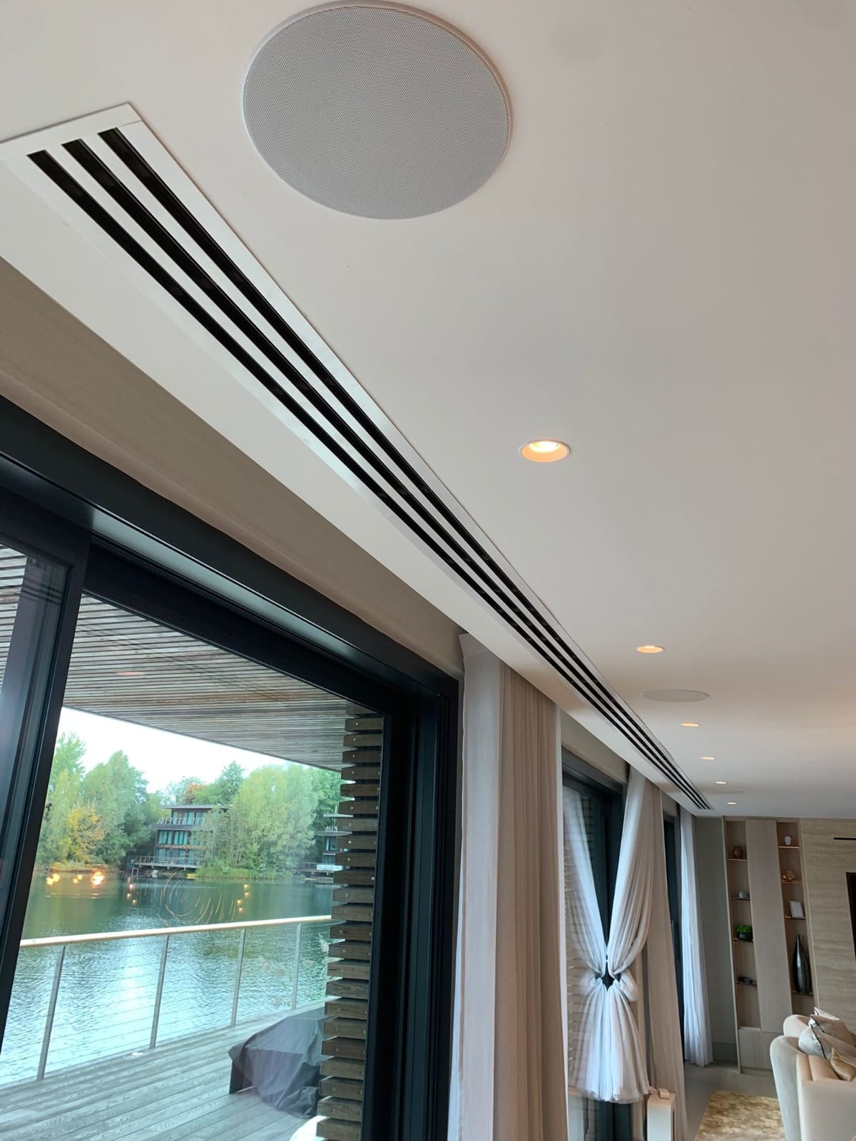 Nowoczesny system wentylacji liniowej z trzema szczelinami, wkomponowany w sufit nad oknem z widokiem na jezioro. Okrągły nawiewnik sufitowy w tle.